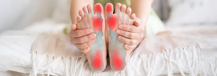 Chiropractic Quincy MA Foot Pain A person holding their feet while sitting on a bed. Their feet are highlighted red.
